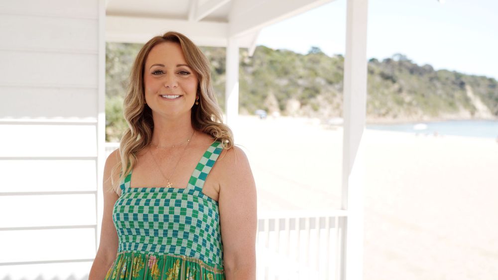Woman in turquoise checkered dress standing on white porch with coastal view.