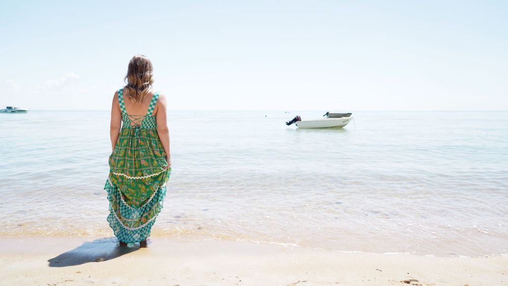 Person in flowing green dress standing at water's edge with boats in distance.