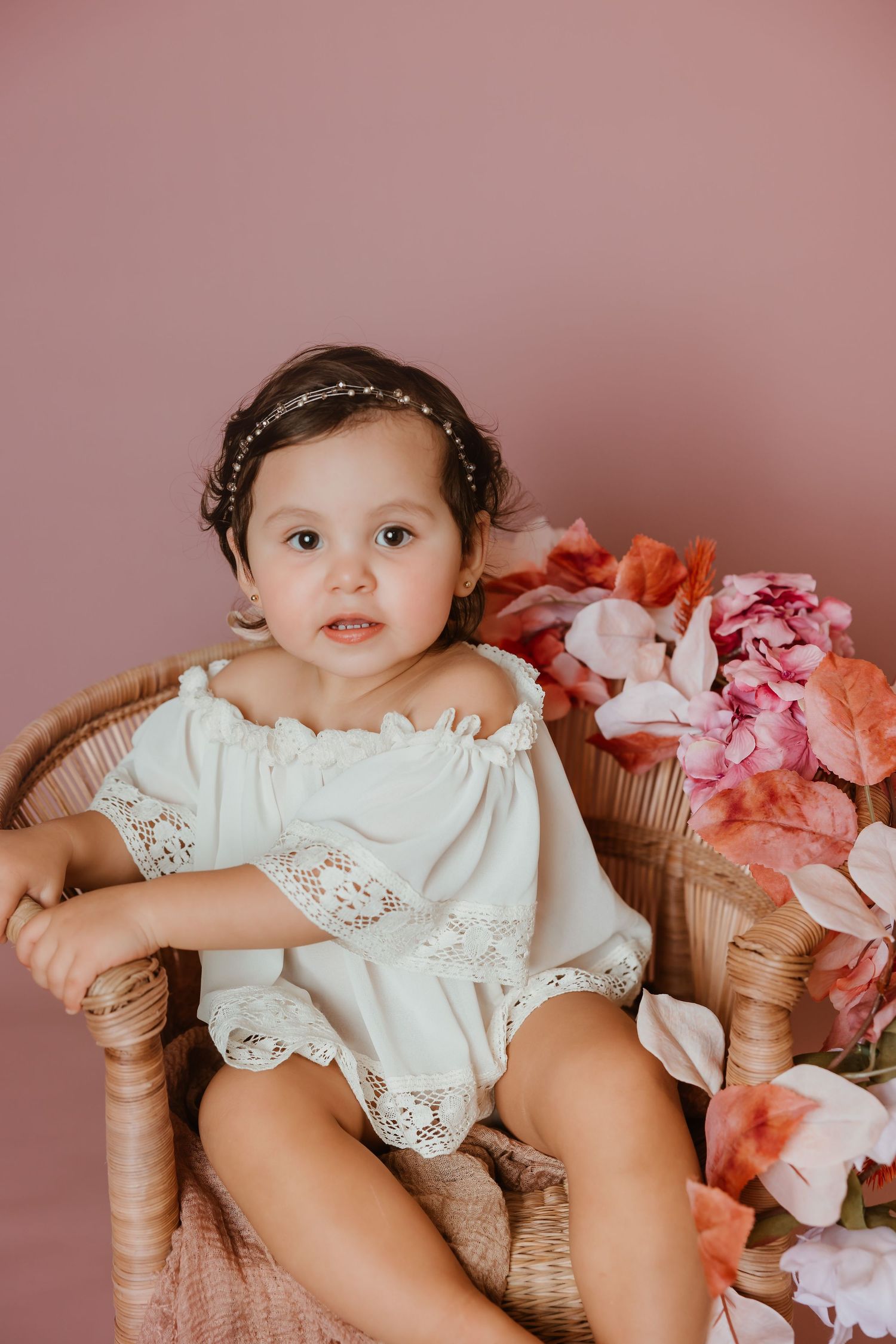 Sweet portrait of toddler in white lace dress on wicker chair with pink flowers.