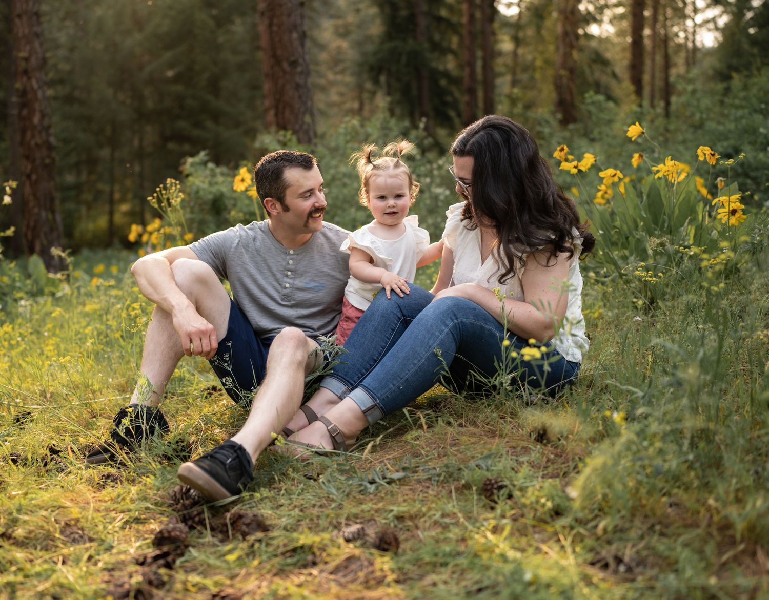 A family relaxes together in a meadow of yellow wildflowers surrounded by tall pine trees.