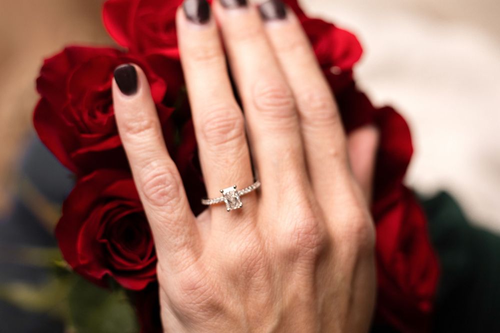 Close-up of a hand wearing an engagement ring with red roses in the background.