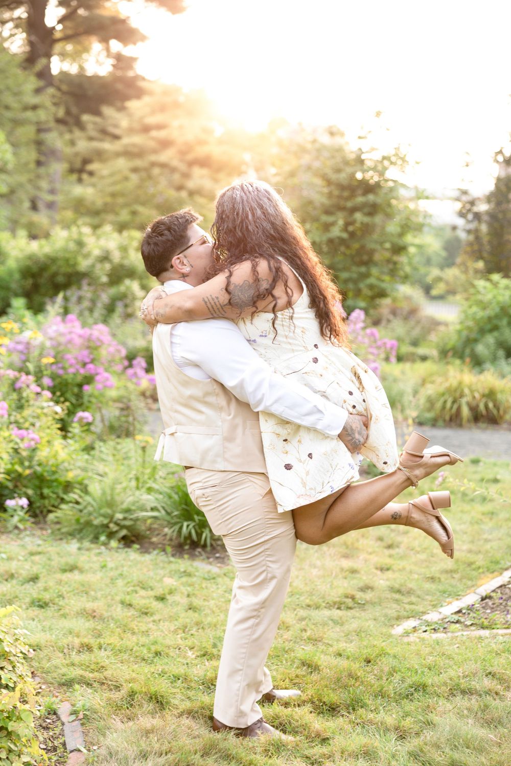 Joyful couple in white outfits sharing a playful moment in a blooming garden at sunset.