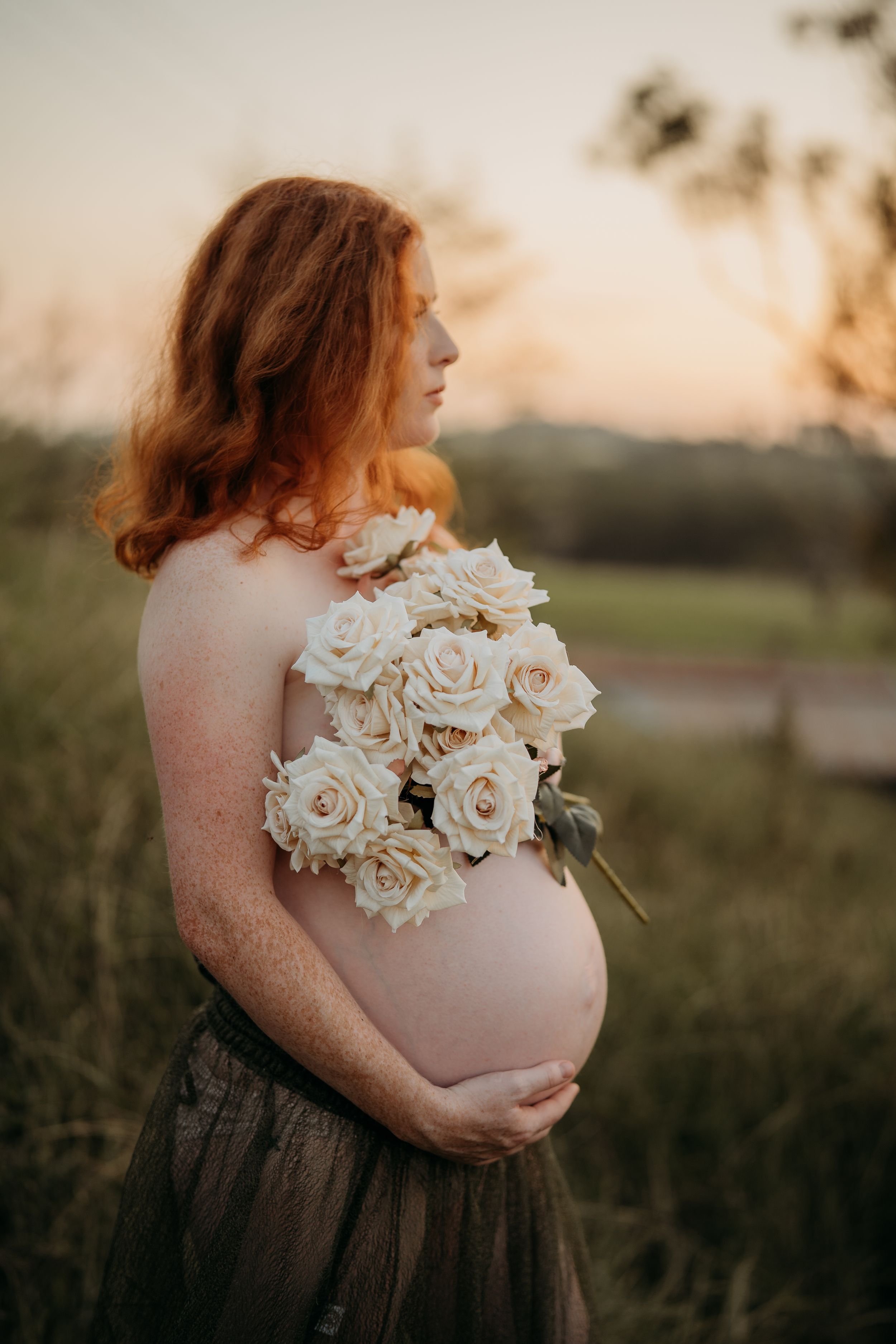 Side profile of a red-haired pregnant woman holding white roses against her bare baby bump at dusk.