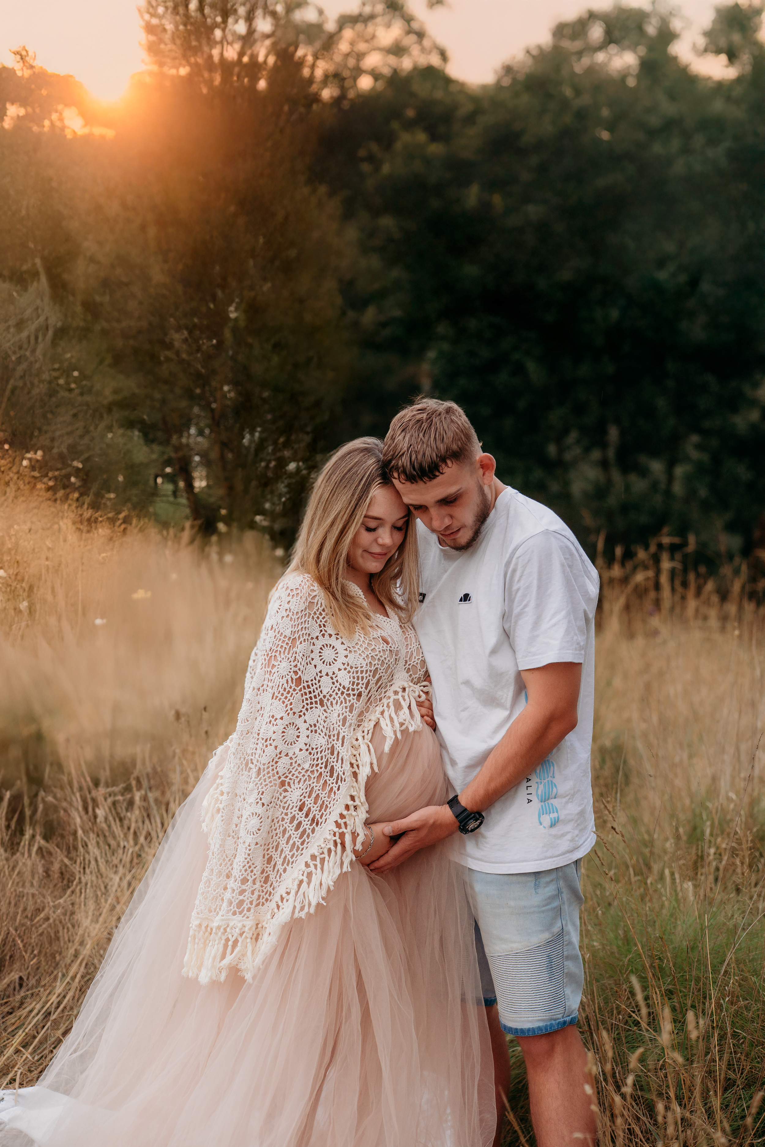 A couple embraces at sunset in a field, the woman wearing a blush tulle skirt and lace shawl.