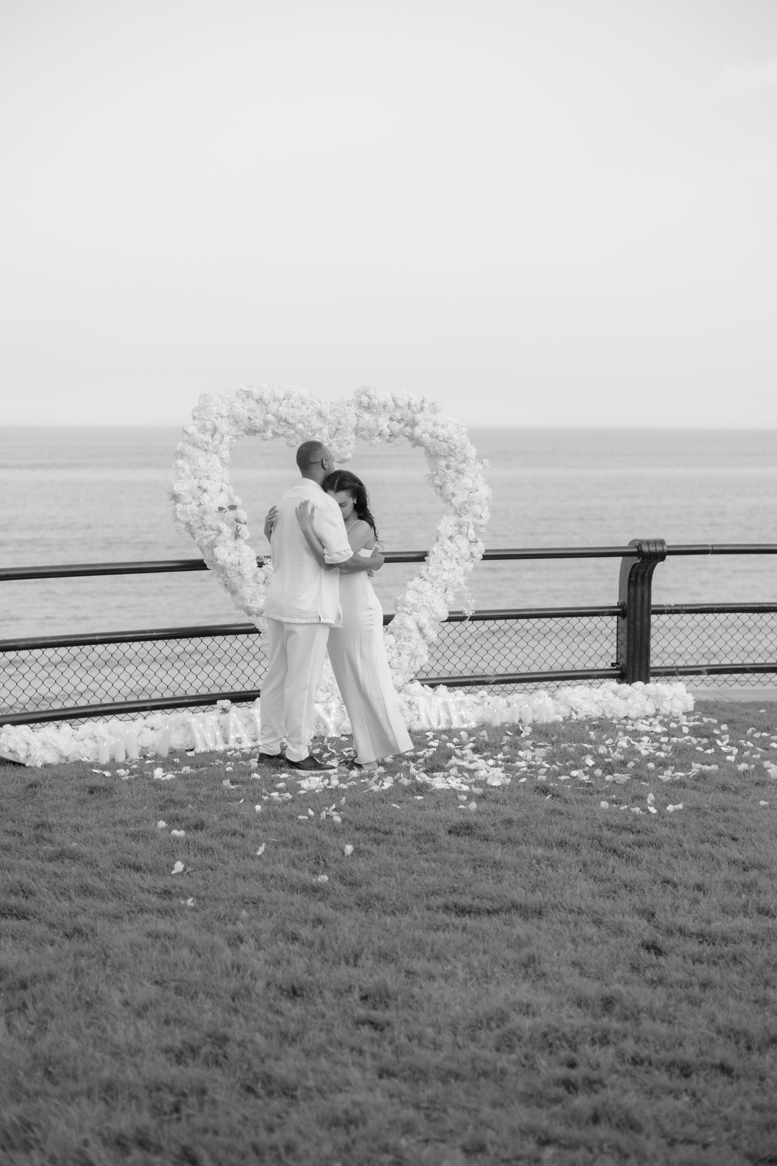 A romantic wedding ceremony under a white floral arch overlooking the water in black and white.