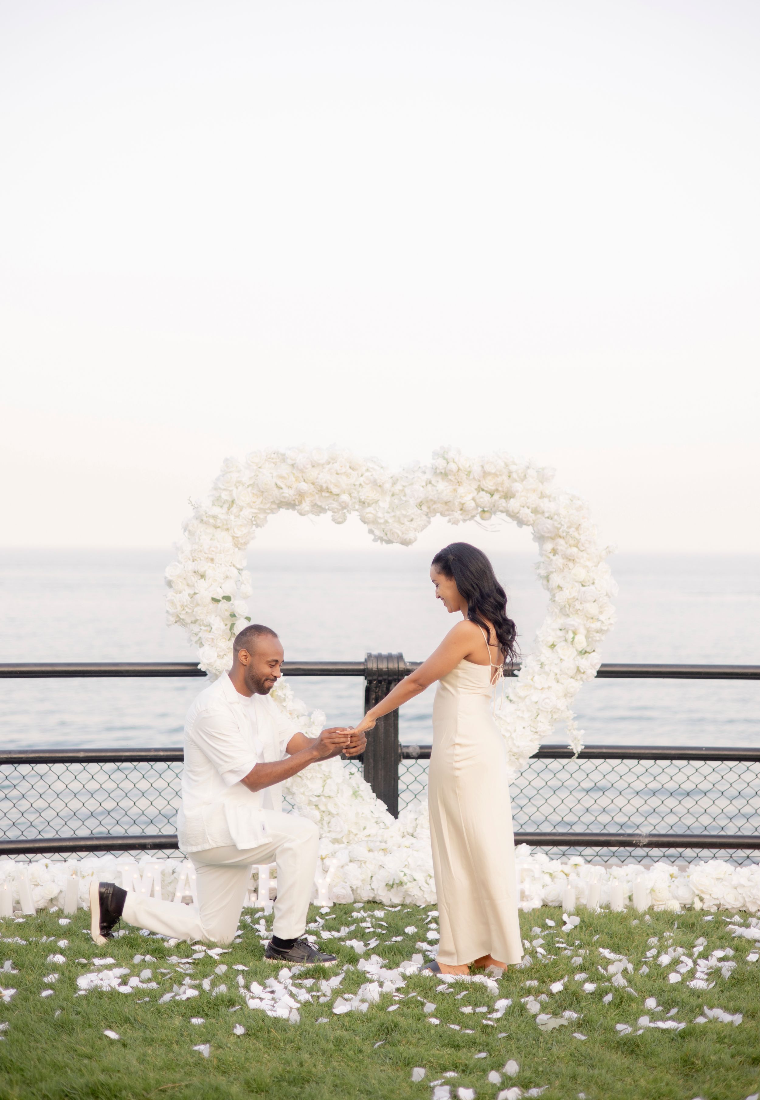 A marriage proposal at sunset under an elegant white floral arch by the waterfront.