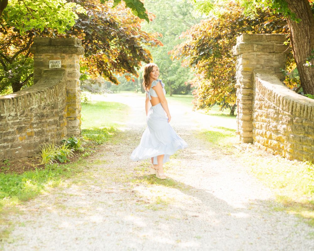 Senior figure in white dress twirls through stone archway on sunlit garden path surrounded by autumn foliage. Kentucky senior photographer