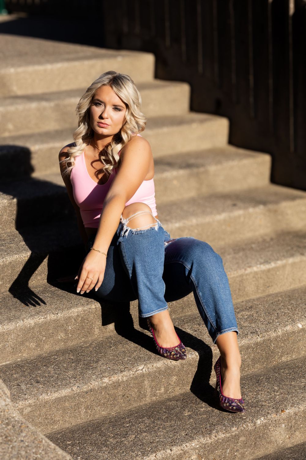 Senior in ripped jeans and sleeveless top poses on concrete stairs in dramatic lighting with shadows. Kentucky senior photographer
