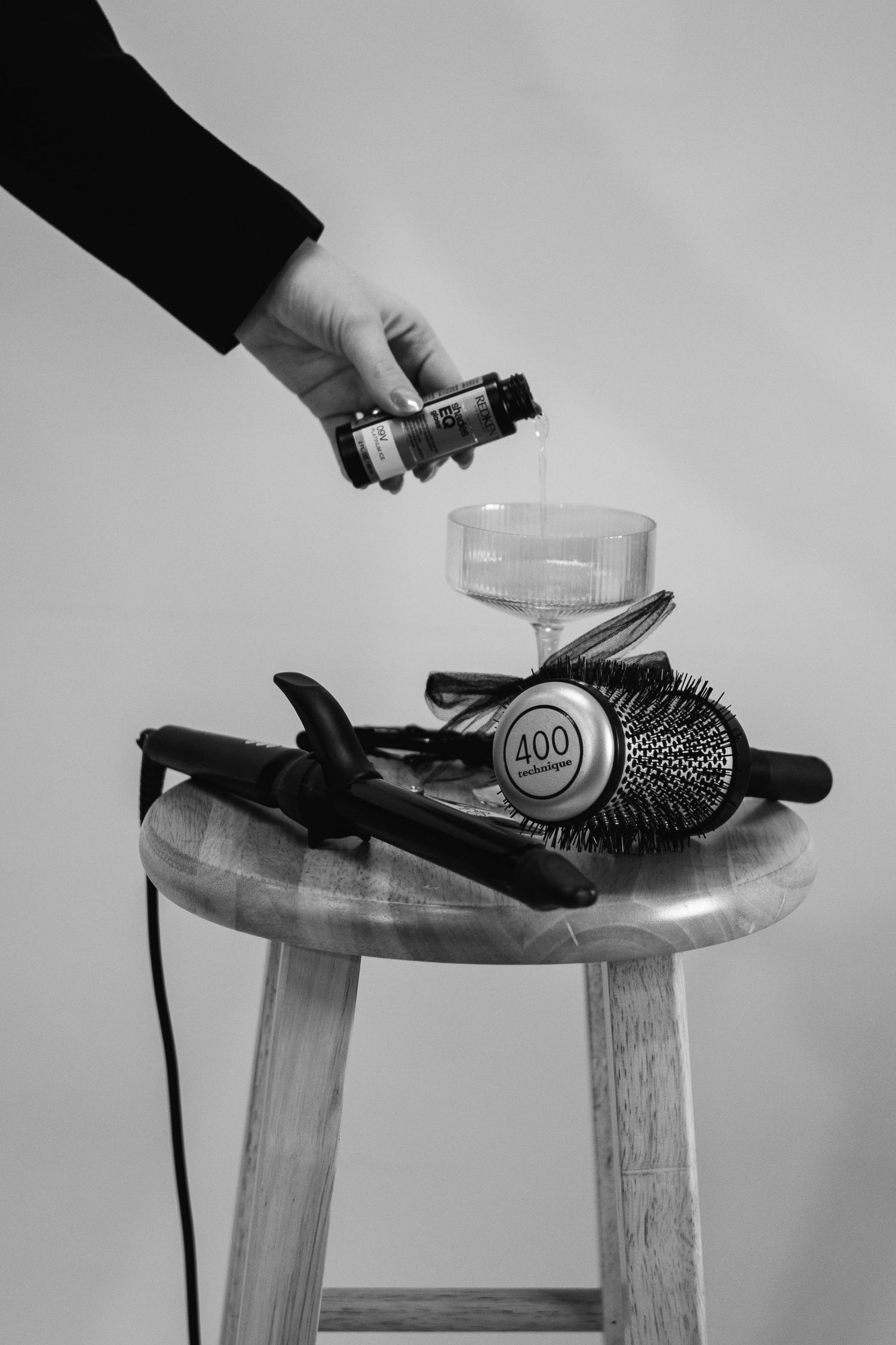Hand pouring hair serum over styling tools including flat iron, round brush, and curling wand on wooden stool.