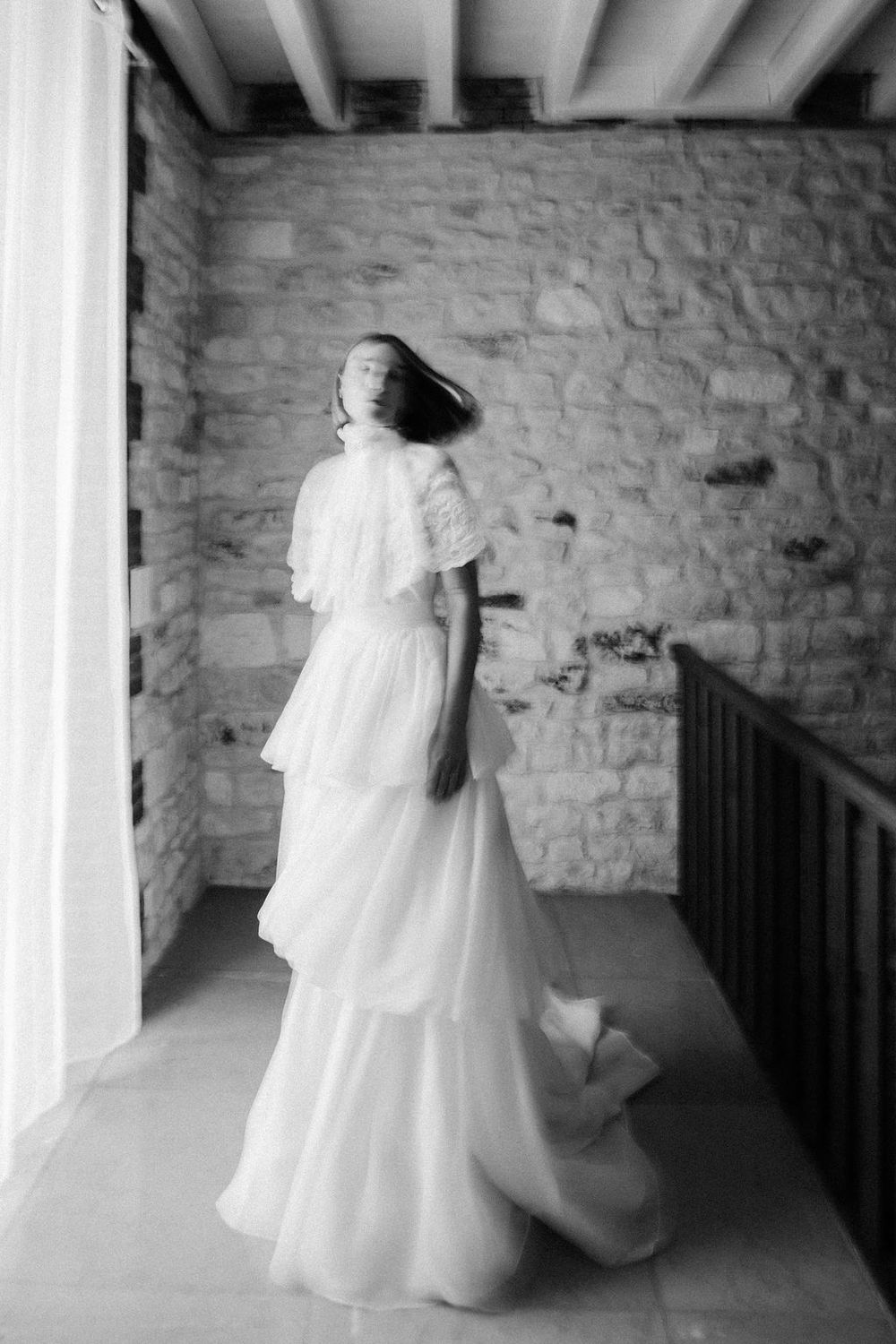 Black and white photo of bride in tiered white gown posing gracefully on a rustic stone balcony.
