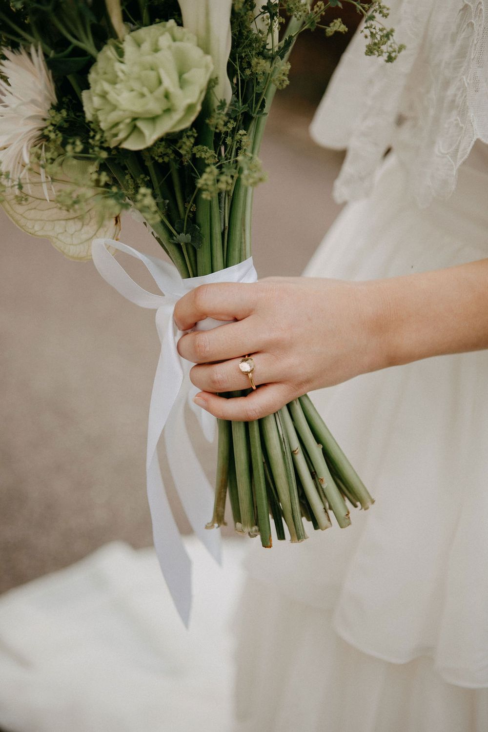 Close-up of bride holding a green and white bouquet tied with white ribbon, wearing a gold ring.