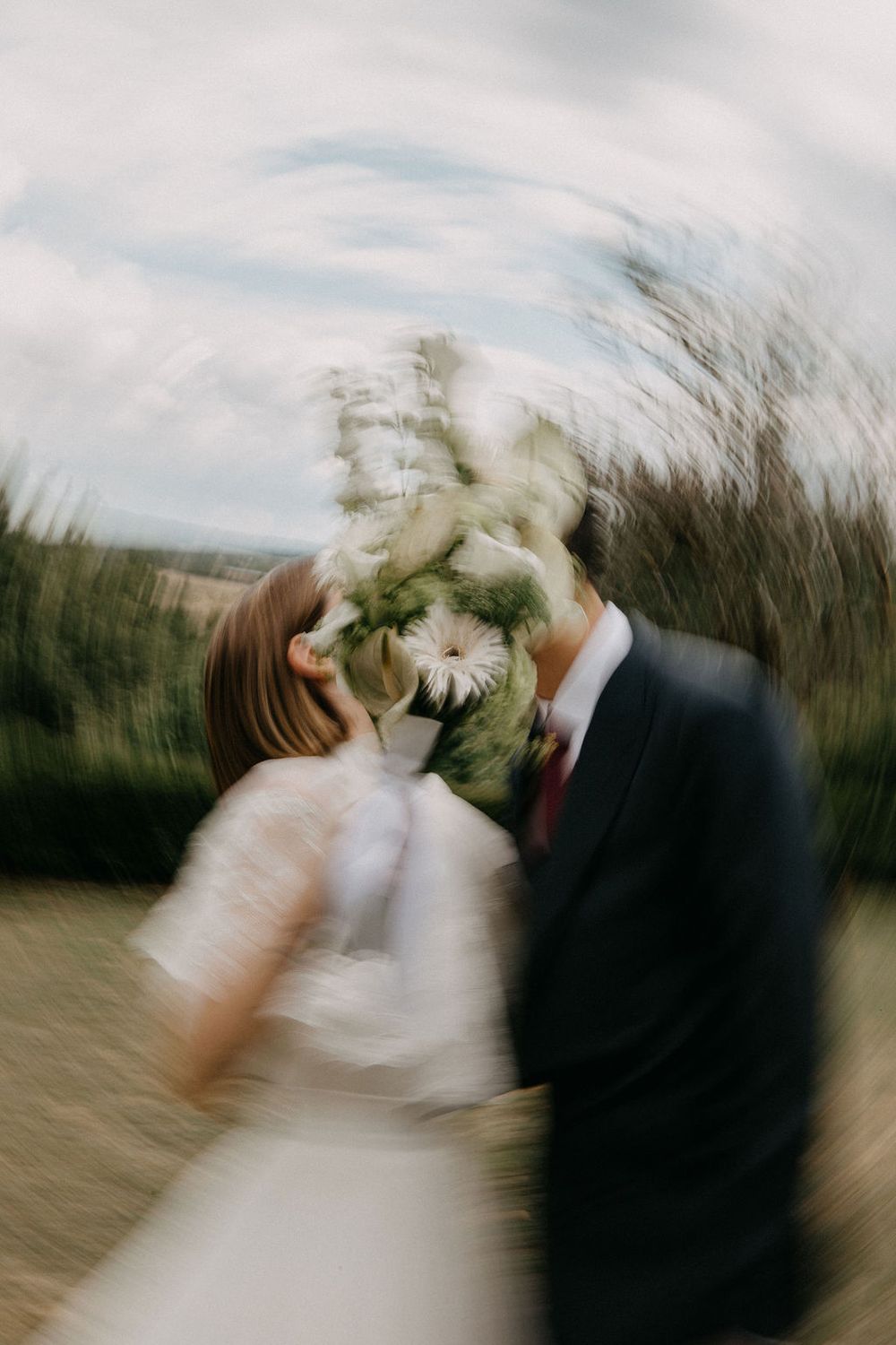 Artistic radial blur photo of bride and groom kissing behind a white and green wedding bouquet outdoors.