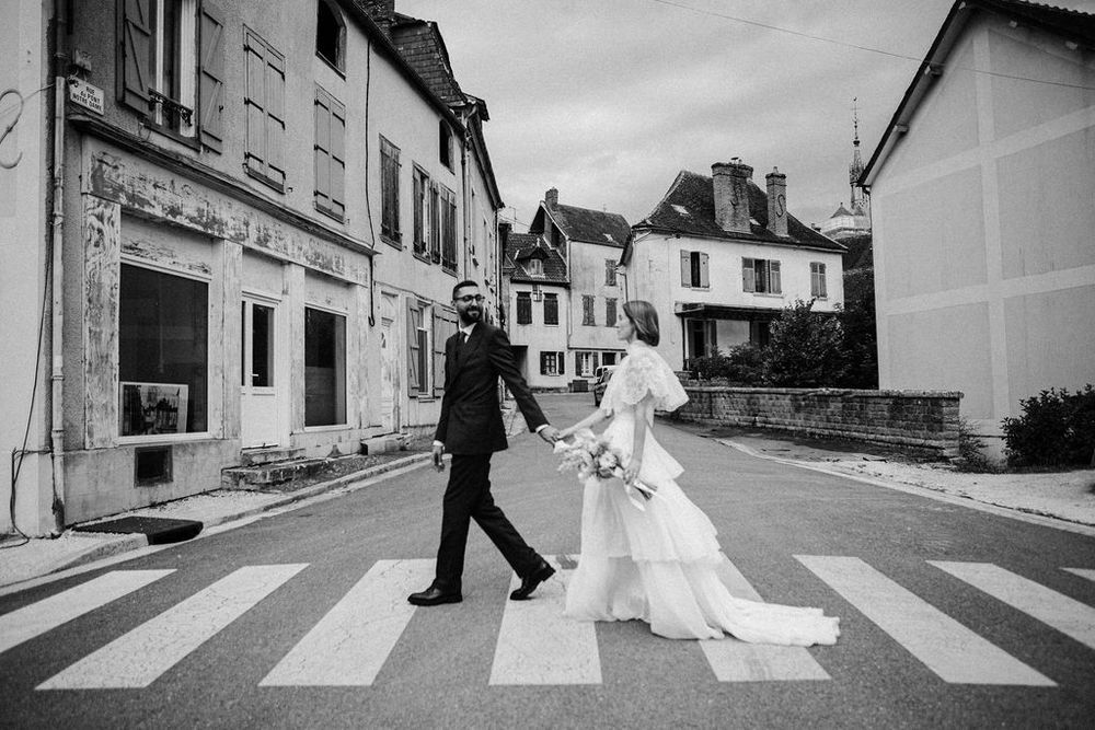 Black and white photo of bride and groom crossing a zebra crossing in a charming French village street.
