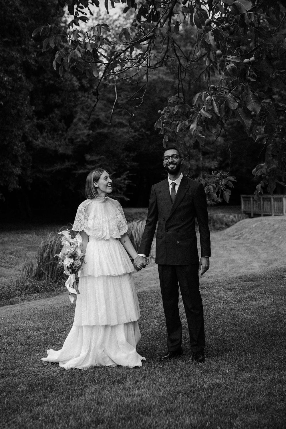 Black and white portrait of smiling bride in tiered lace gown and groom holding hands in a garden.