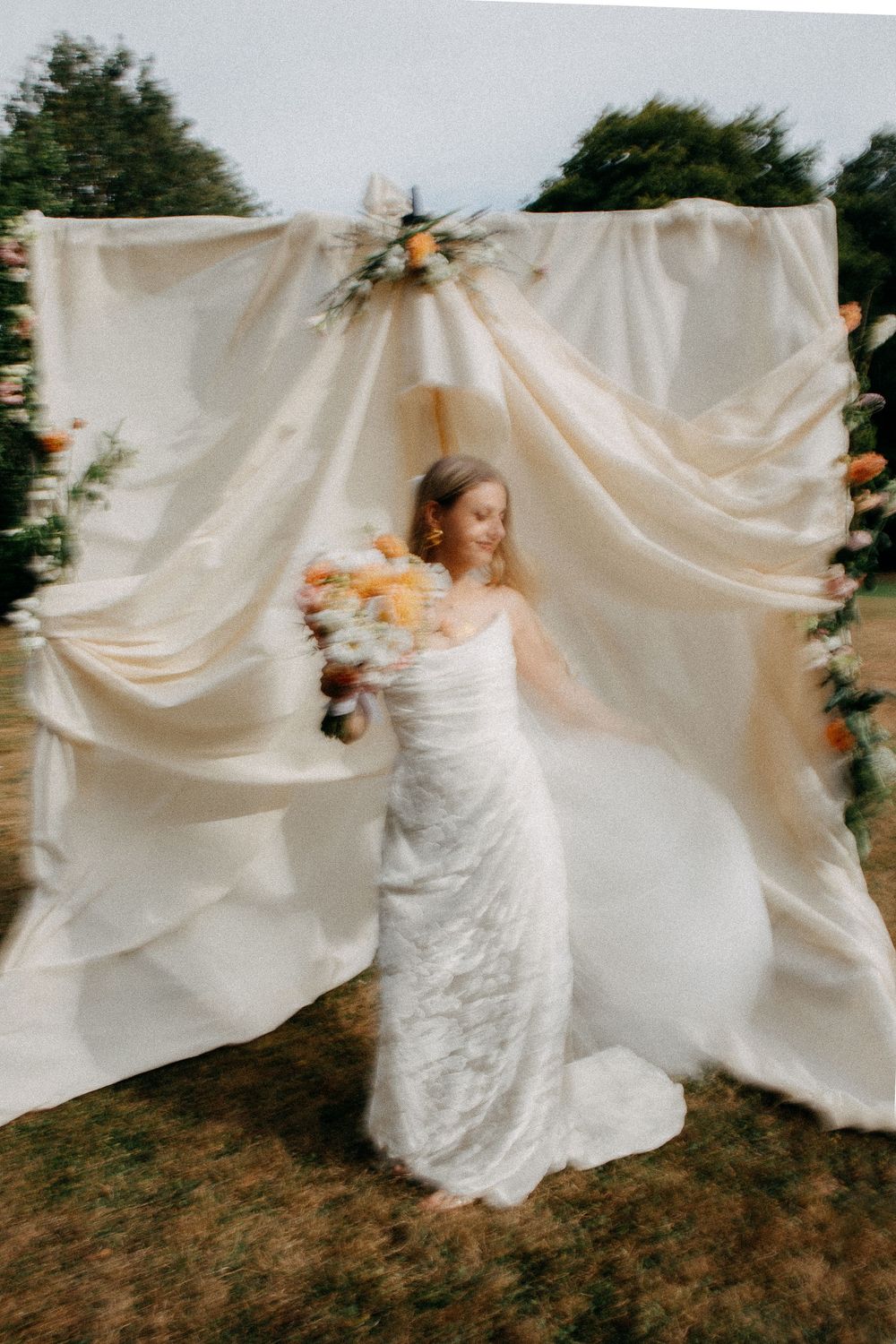 Bride in white lace gown holds colorful bouquet before draped fabric backdrop with floral accents outdoors.