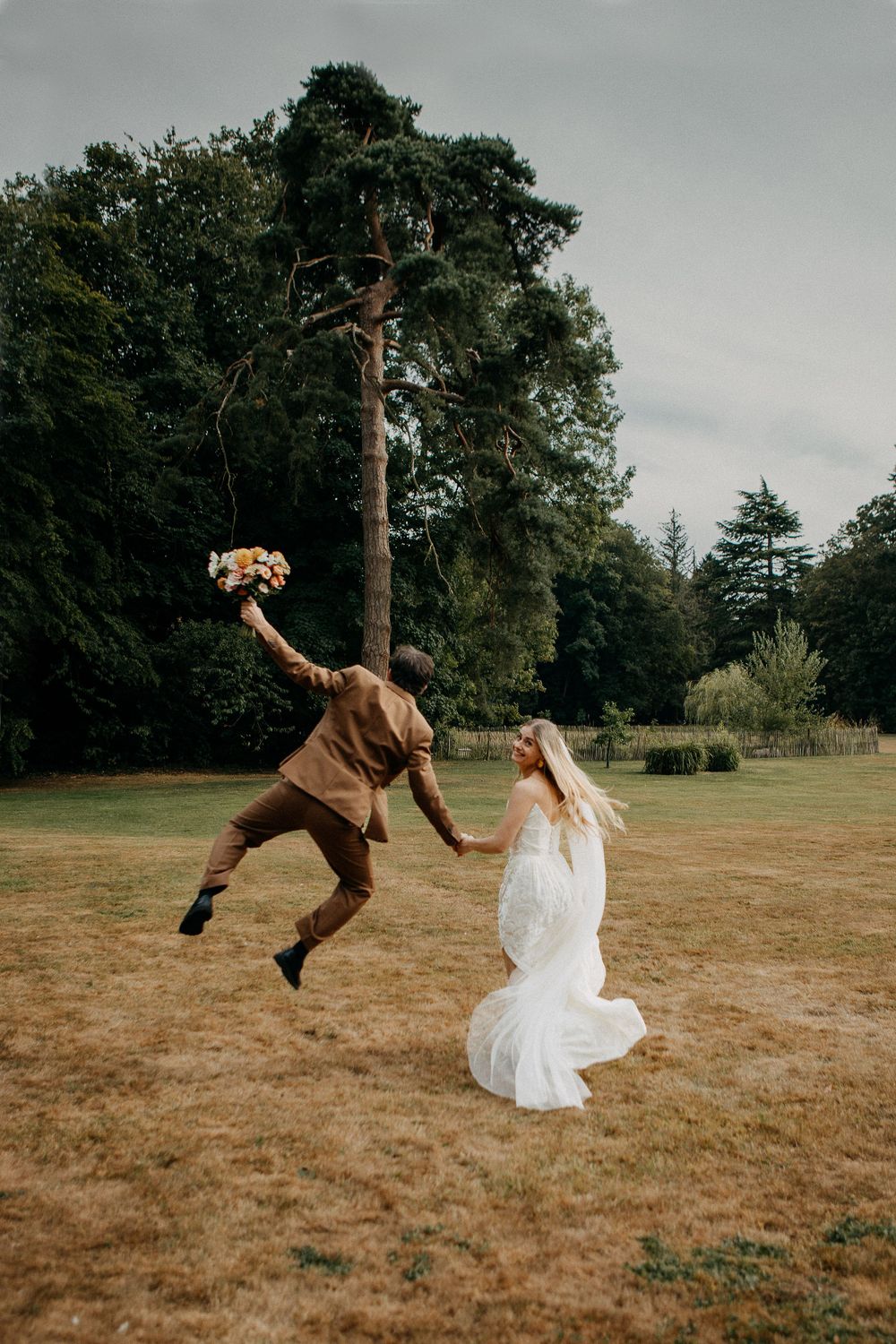 Joyful groom jumping with bouquet while holding bride's hand in white gown on lush green lawn.