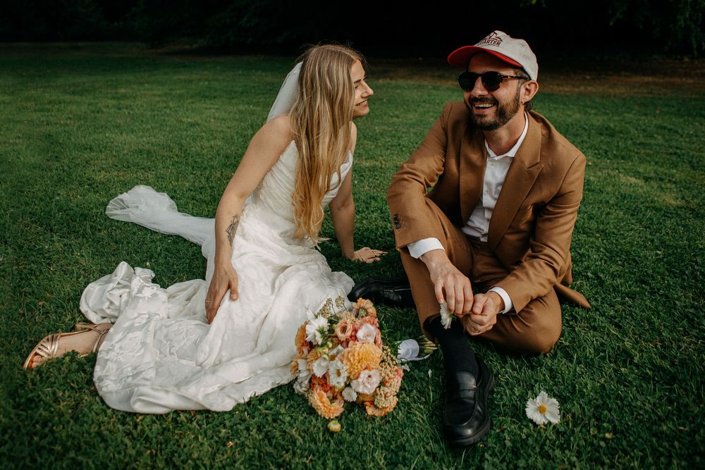 Bride and groom sitting on green grass, laughing together, bouquet nearby, groom wearing brown suit and cap.