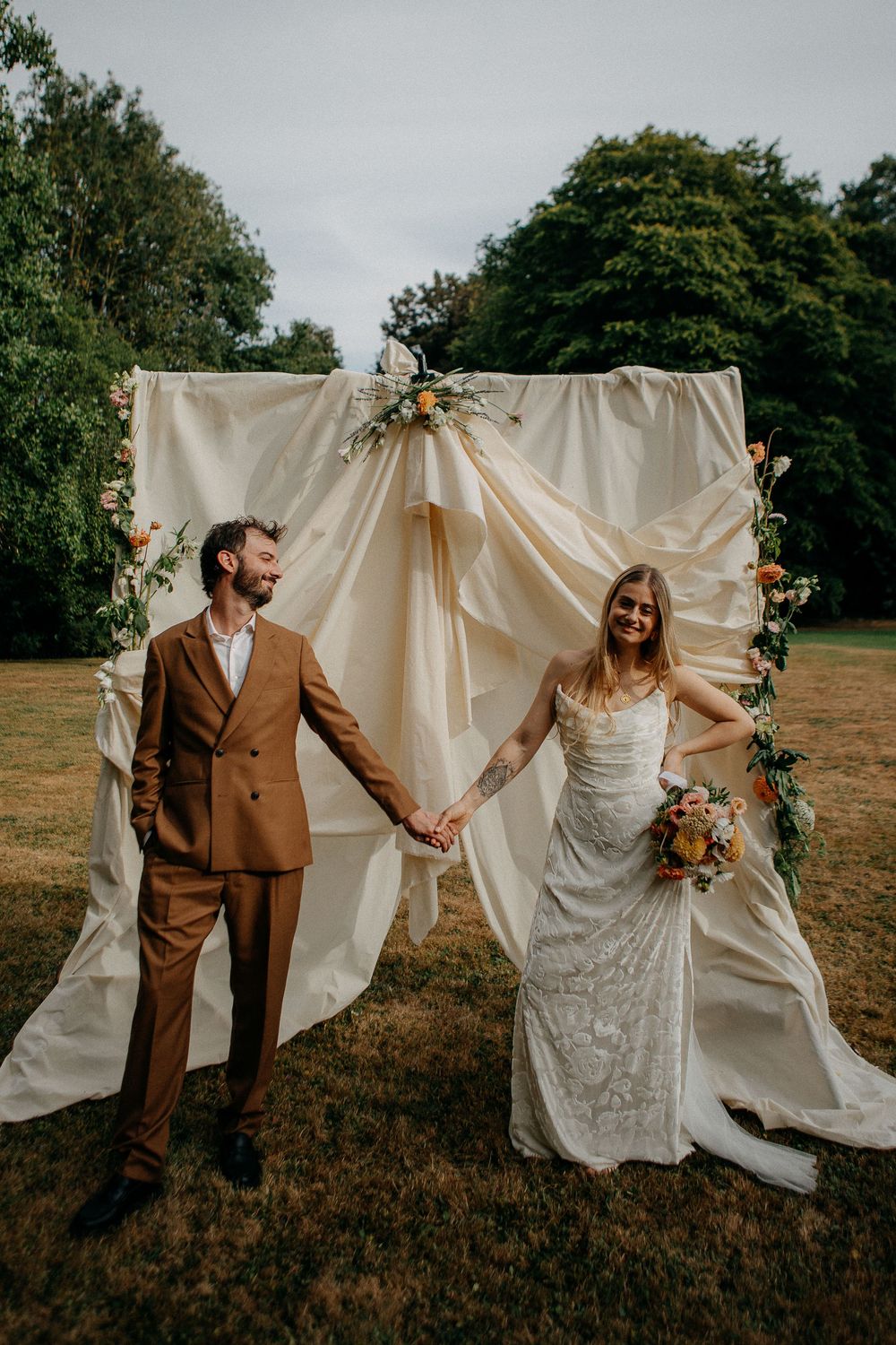 Bride and groom hold hands in front of a flowing cream fabric backdrop with floral accents at an outdoor wedding ceremony.