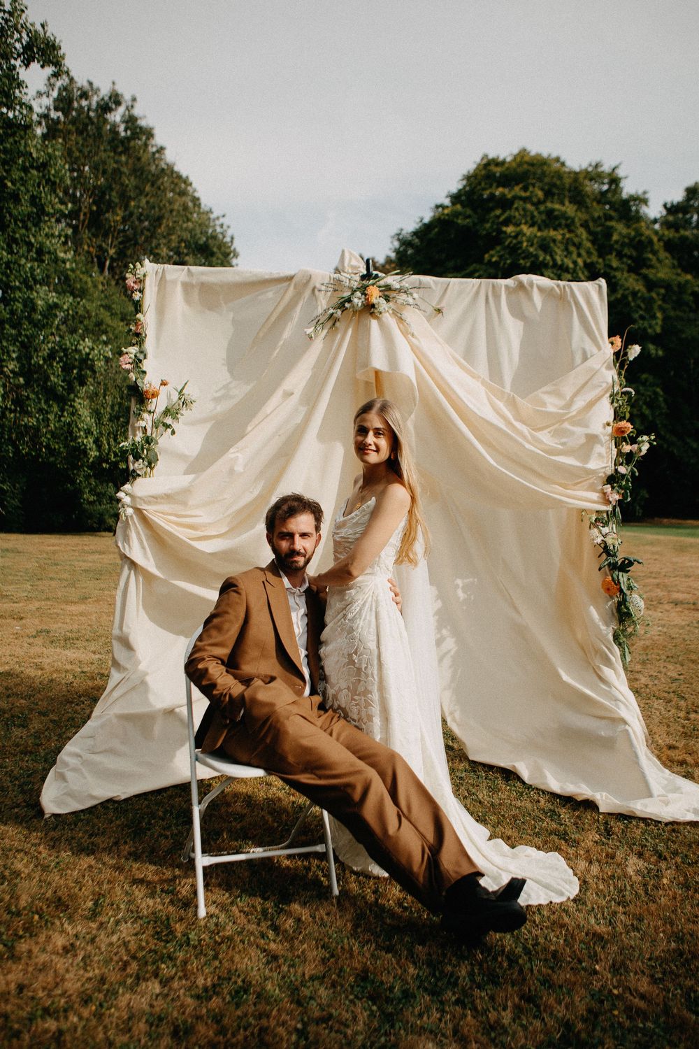 Bride and groom pose before cream draped fabric altar with flowers outdoors on grass at elegant garden wedding.