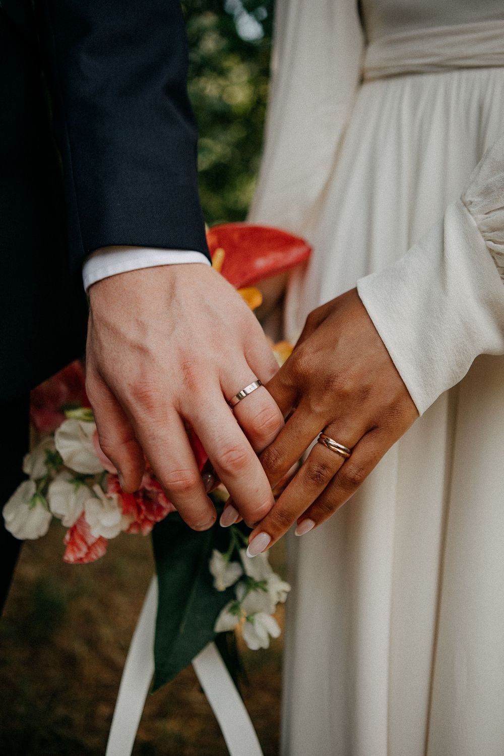 Newlywed couple holding hands showing matching wedding rings, bride in white dress holding colorful bouquet.