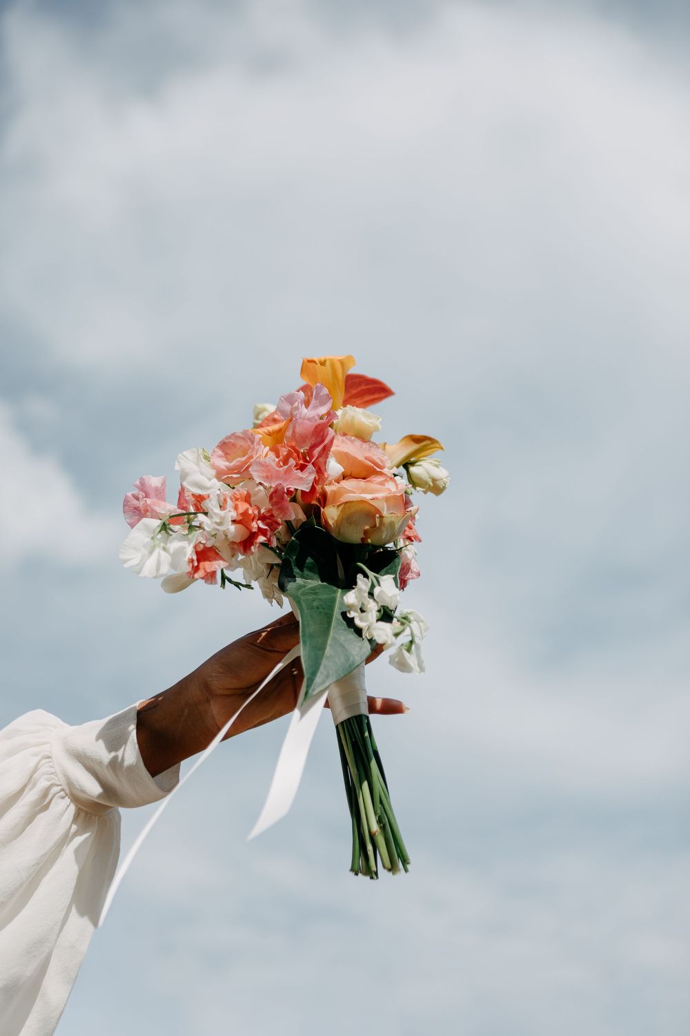 A hand holding a colorful bridal bouquet of pink, orange, and white flowers raised against a cloudy blue sky.
