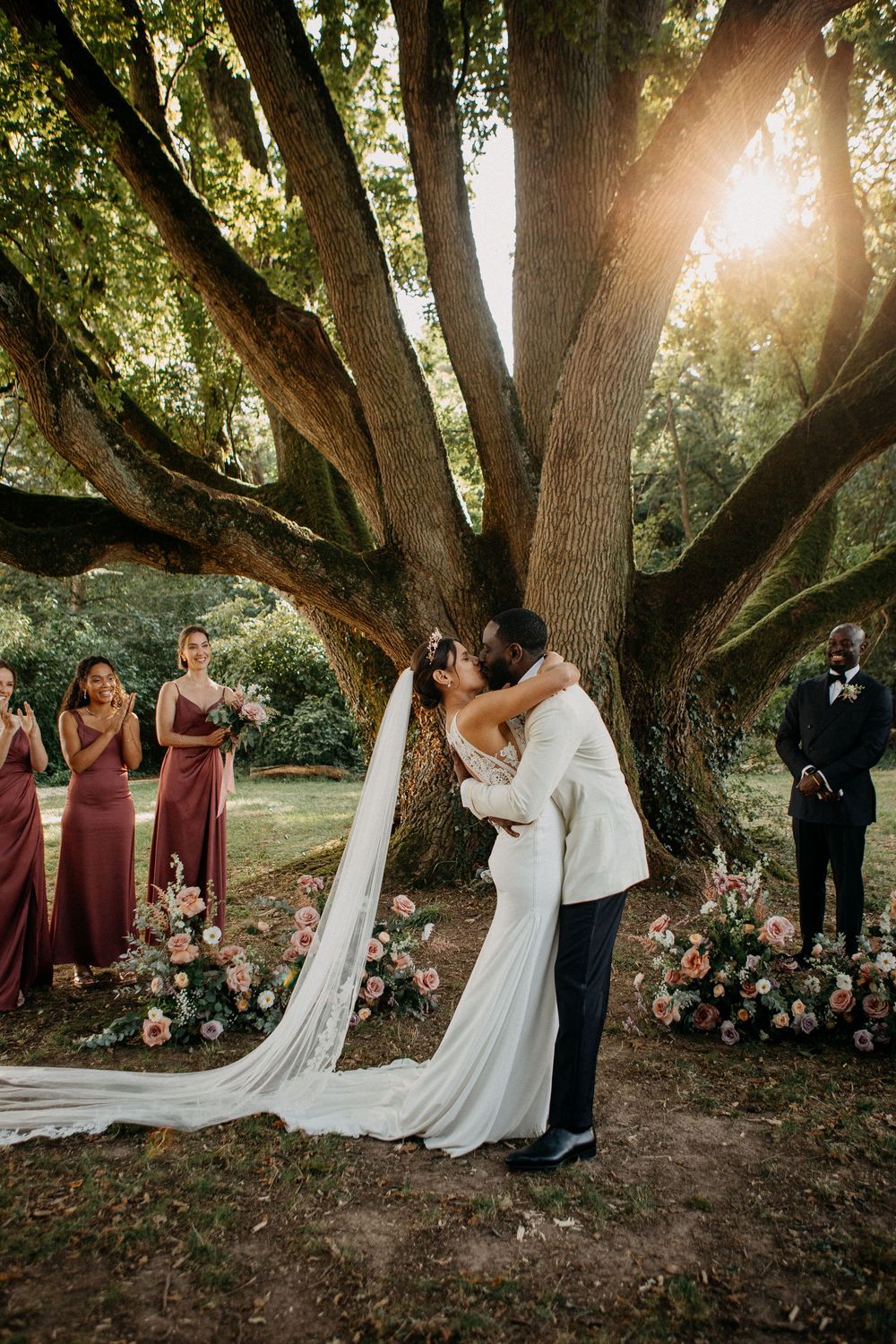 Bride and groom share first kiss under a majestic tree with golden sunlight at outdoor ceremony.