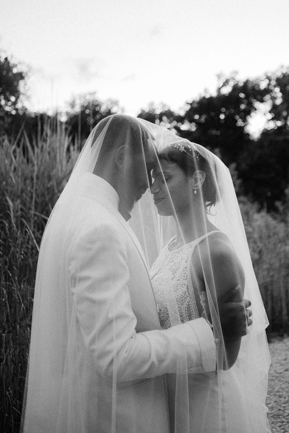 Black and white romantic photo of bride and groom sharing a kiss under a wedding veil outdoors.