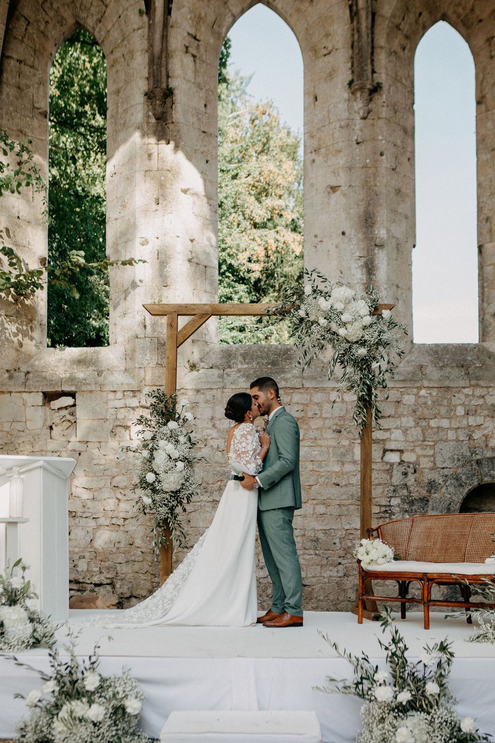 Bride and groom share first kiss at elegant outdoor altar adorned with white floral arrangements in stone ruins.