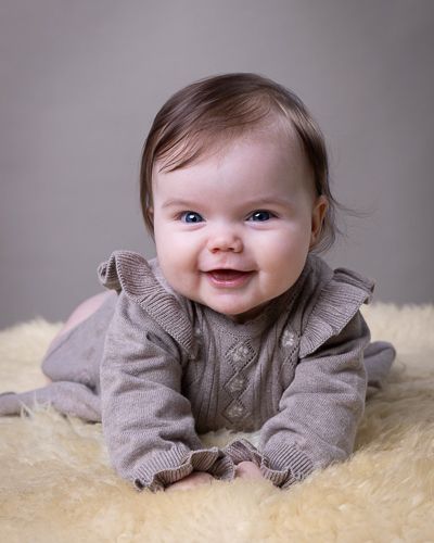 Baby med brun kjole liggende på saueskinn og grå bakgrunn. Babyfotografering i studio på Fjerdingby på Romerike