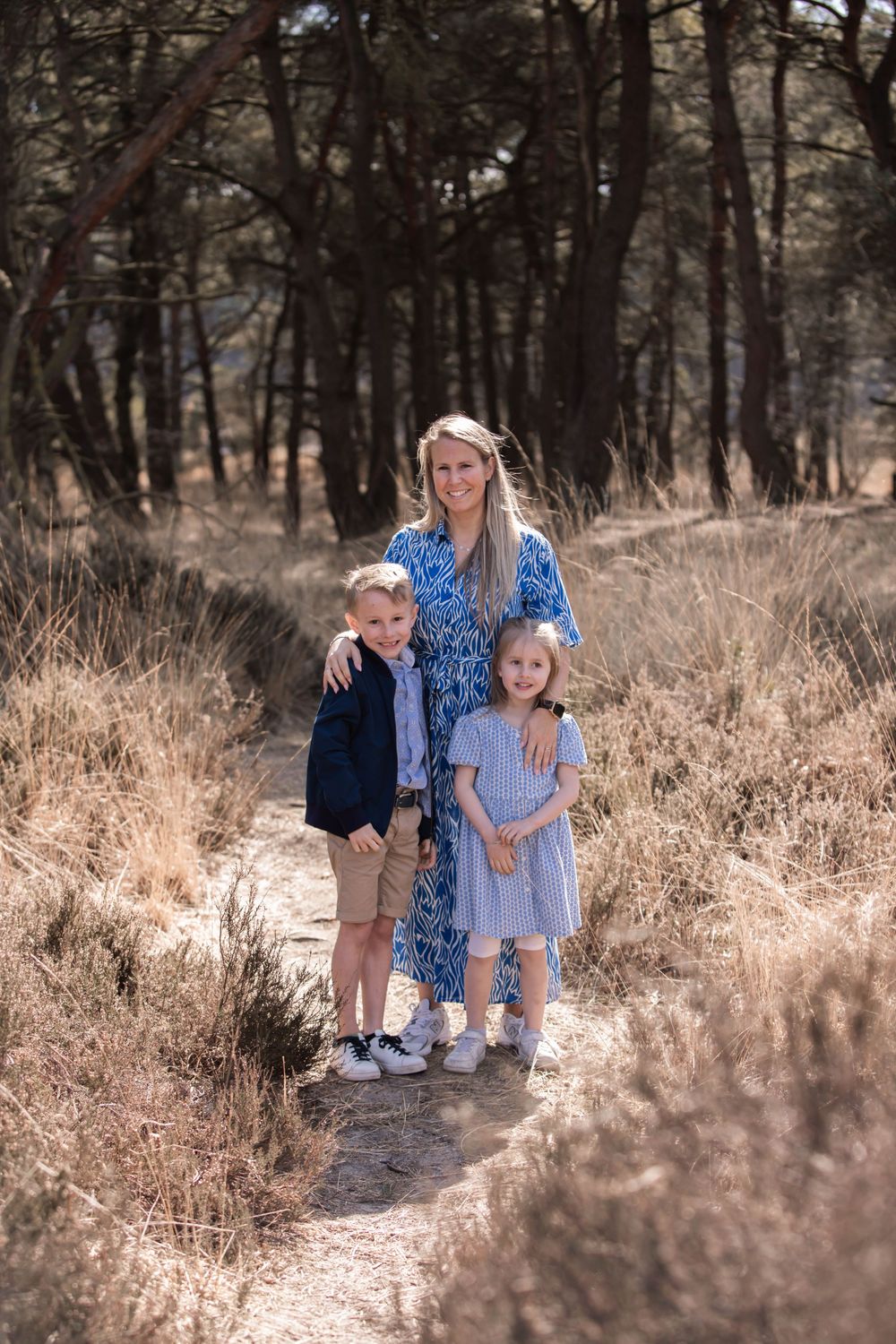 A family wearing blue outfits stands together on a wooded trail surrounded by tall pines and brown grass.