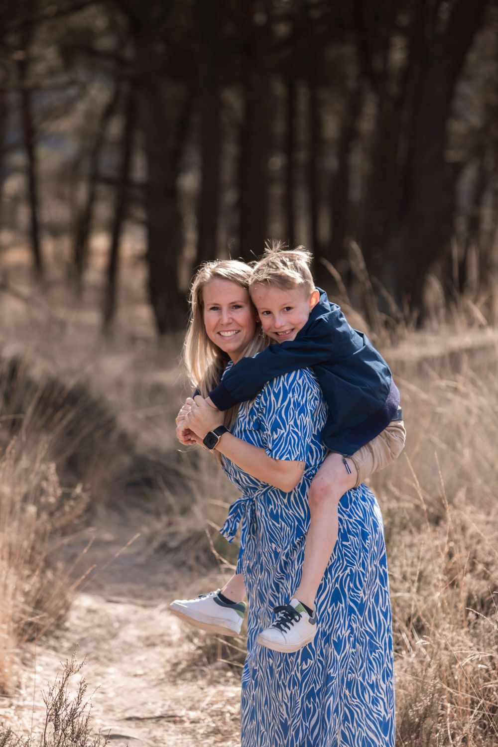 A parent and child share a playful moment in casual outdoor attire against a wooded forest backdrop on a sunny day.
