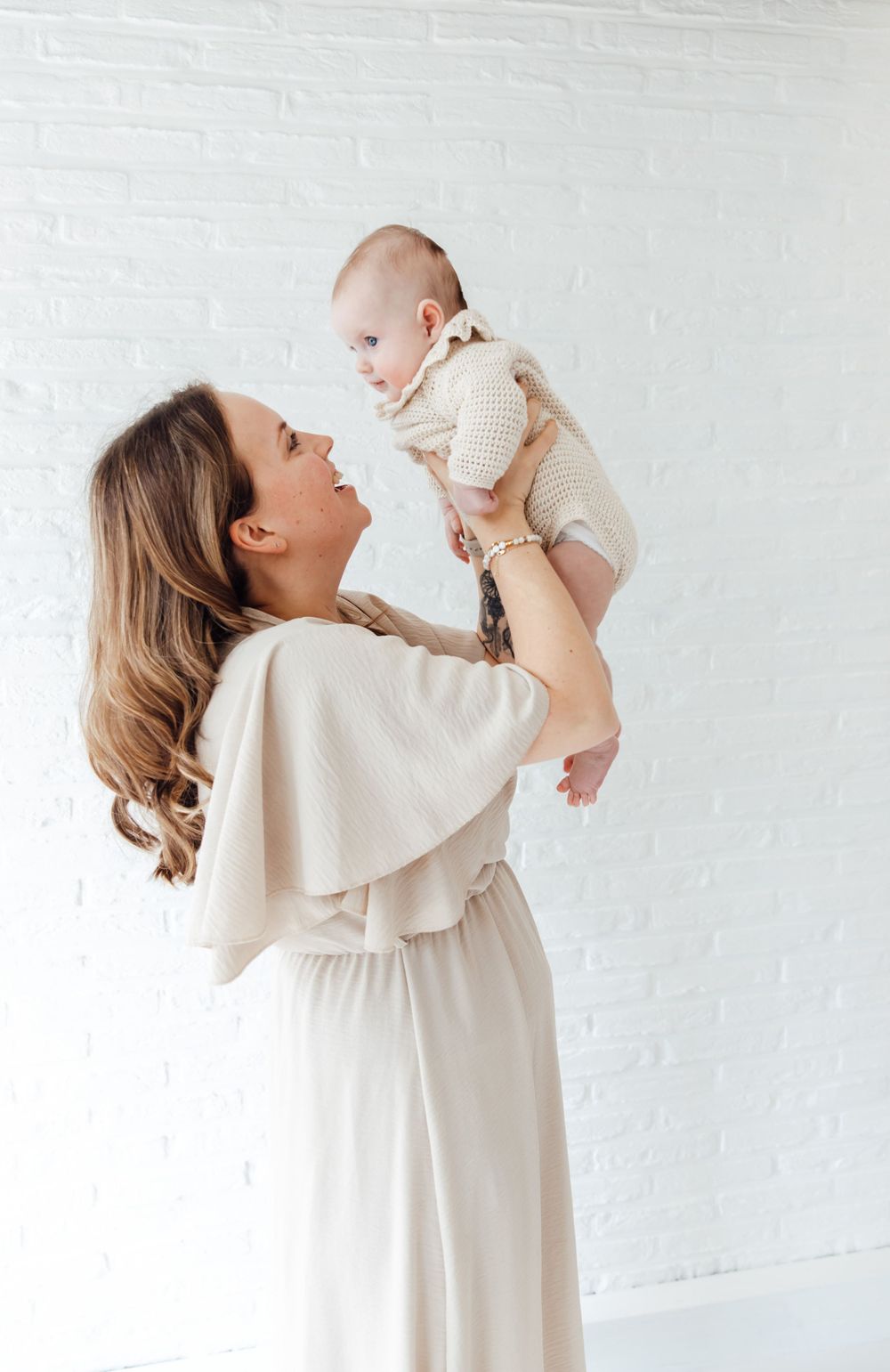 Mother in cream dress lifts infant wearing cream onesie against white wall, sharing tender loving moment.