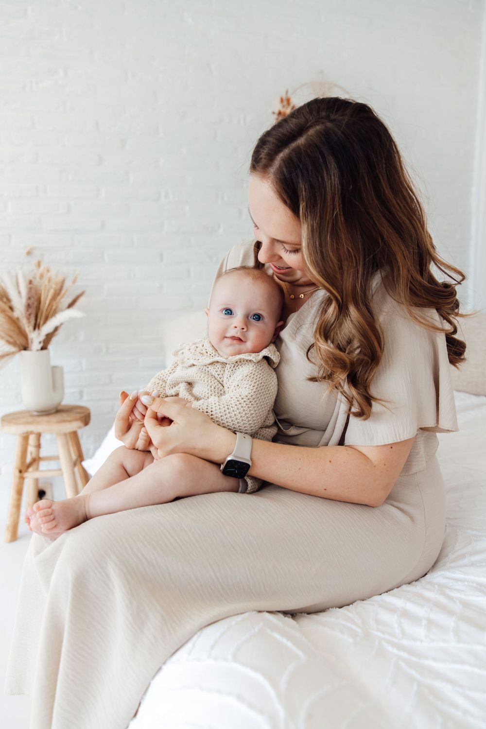 Parent sits on bed with baby in cream sweater, dried flowers in background.