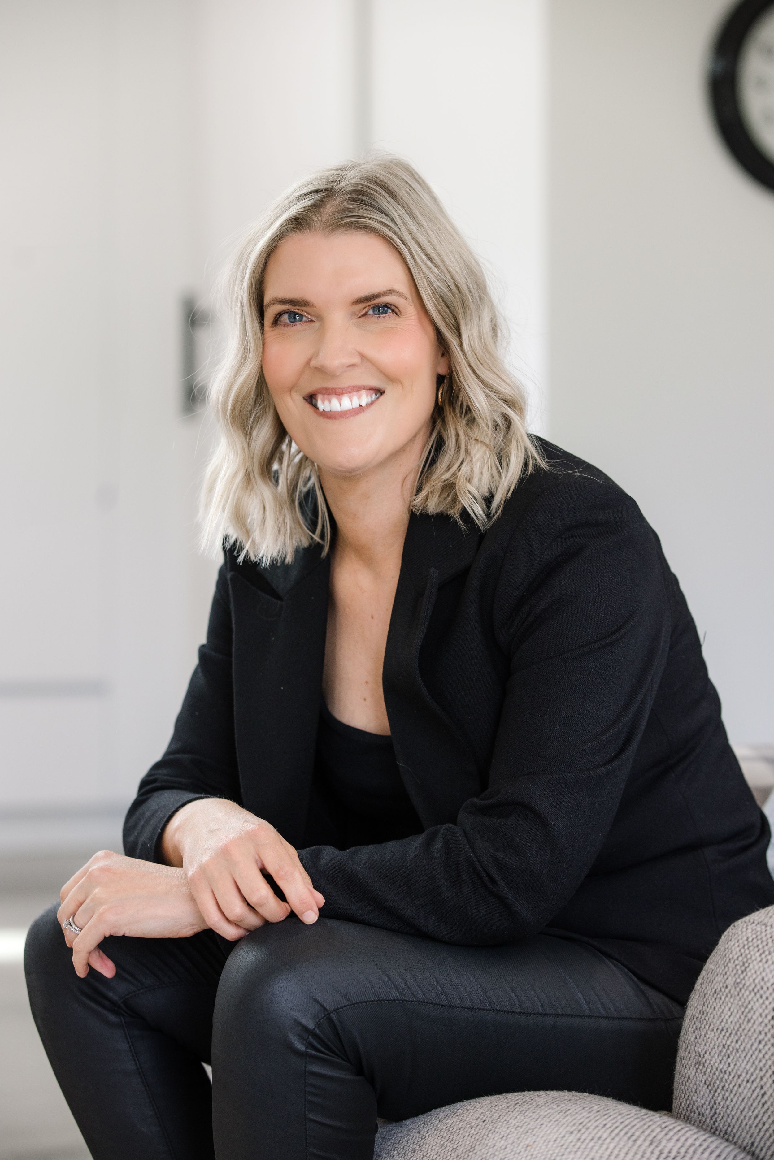 Professional woman in black business attire sits casually on a chair with a warm smile against a light interior background.