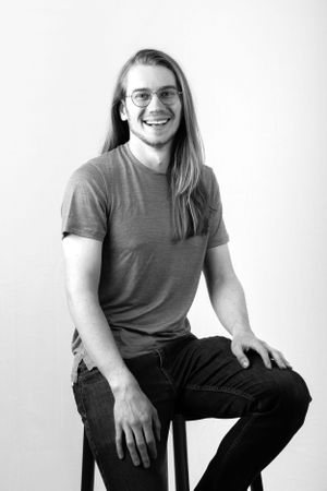 Professional headshot of a person with long hair and glasses wearing a gray t-shirt against a white background.