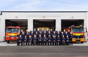 Large group of firefighters in dress uniforms posing outside a fire station with two fire engines.