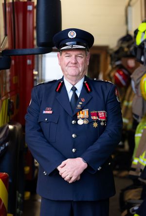 Senior firefighter in dress uniform with medals stands proudly in fire station bay.