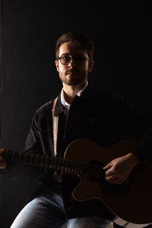 Low key moody portrait of musician in black clothing holding an acoustic guitar against dark background.