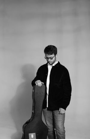 A person in glasses and jacket looking down at a guitar case in a black and white studio portrait.