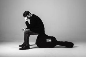 Artistic black and white portrait of person sitting on guitar case in contemplative pose against neutral background.