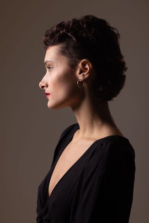 Profile view of a person with dark curly hair wearing a black dress against a neutral brown background in elegant portrait style.
