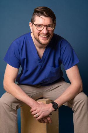 Healthcare professional in blue scrubs seated on wooden stool against blue background.