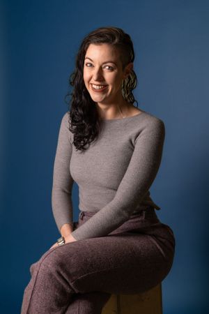 Professional portrait against blue background featuring grey sweater and casual pose.