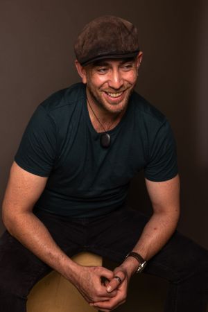 Casual portrait of person wearing flat cap and black t-shirt seated on stool.