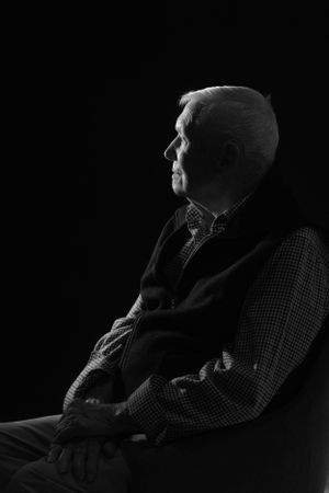 Black and white profile portrait of an elderly person sitting in contemplation against dark background.