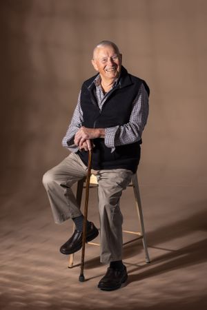 Portrait of a senior person in a black vest and plaid shirt seated on a stool with a walking cane.