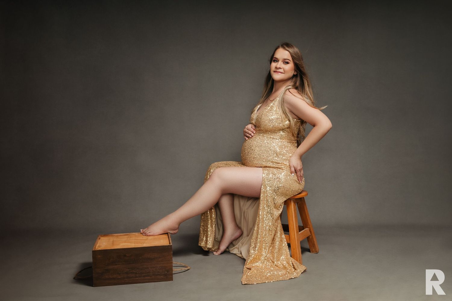 Maternity portrait of woman in gold sequin dress seated on wooden stool.