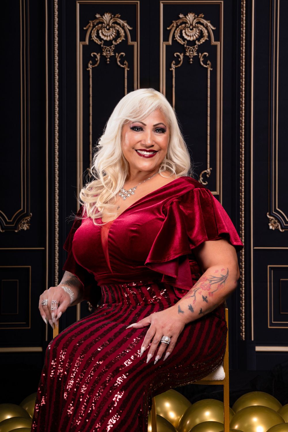 Glamorous portrait against ornate black and gold backdrop with red velvet gown and golden balloons.