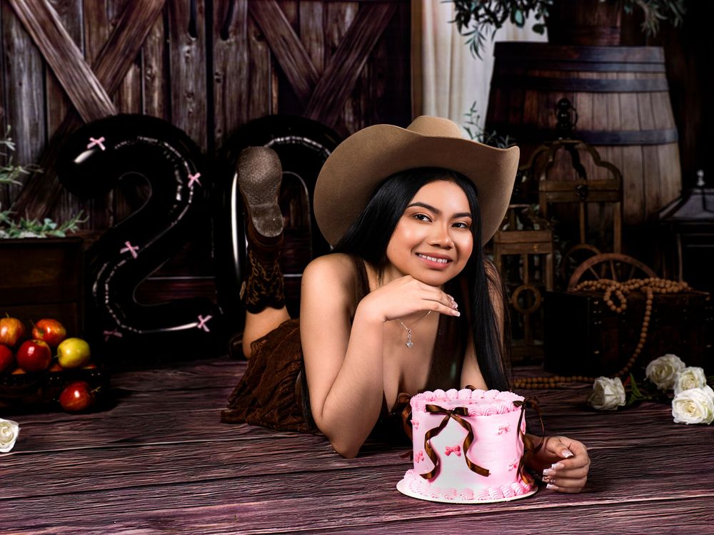 Young woman in cowboy hat smiling beside a pink birthday cake with number decorations in a rustic barn setting.