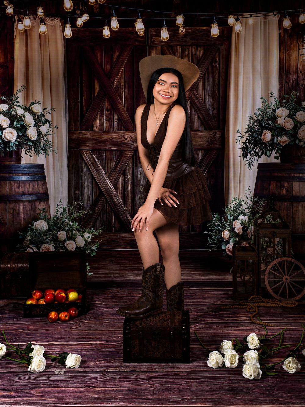 Young woman in brown dress, cowboy hat, and boots posing in rustic barn setting with flowers and string lights.